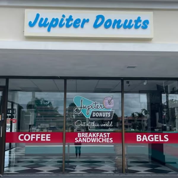 Exterior view of Jupiter Donuts storefront with a blue sign and window decals for coffee, breakfast sandwiches, and bagels.