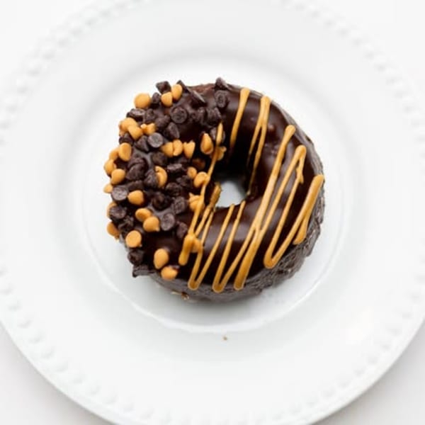 A chocolate donut with chocolate and peanut butter chips, drizzled with peanut butter, on a white plate.